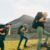 Three girls are rooming in front of a mountain with waering fjallraven trousers and shirts and also with backpacks on their backs. 