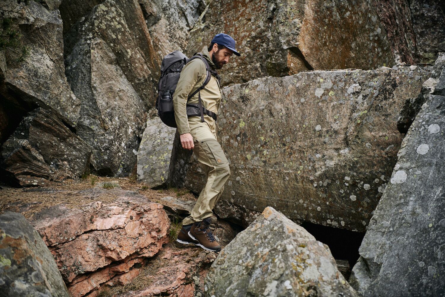 one man is trekking in the rocks wearing dark navy fjallraven t-shirt, dark navy abisko cap and a brown trouser with a large trekking backpack on his back.