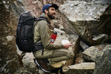 one man is sitting in the middile of mountains during trekking with fjallraven backpack on his back and a branded cap on his head.