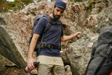 one man is trekking in the rocks wearing dark navy fjallraven t-shirt, dark navy abisko cap and a brown trouser with a large trekking backpack on his back and a small water bottle in his hand.