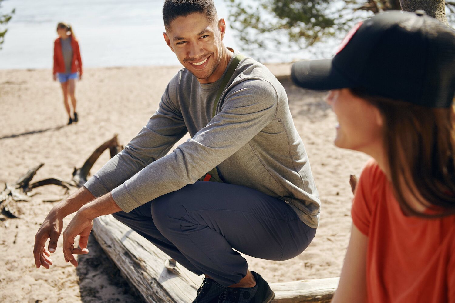 one girl wearing black fjallraven cap with red fox logo and red t-shirt is sitting with a guy on beach.  and one more girl is coming towards them from backside.